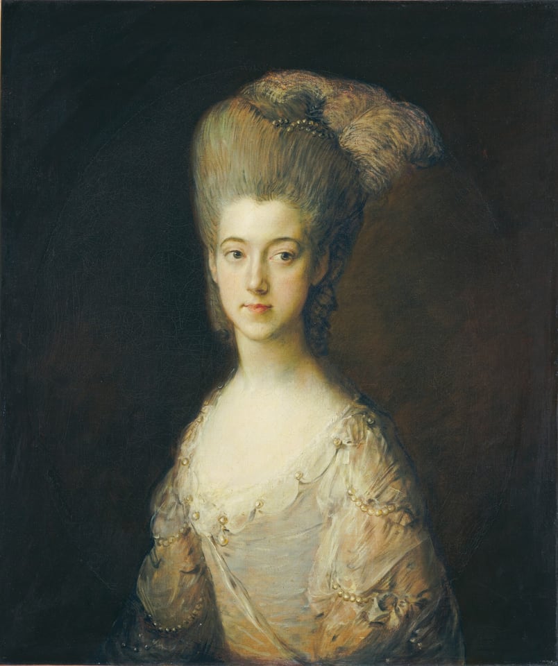 Shown from the waist up, a young, pale-skinned woman with her gray hair piled high atop her head is lit starkly against a dark background in this vertical portrait painting. The woman’s shoulders are angled to our left. Her face is square to us, but she looks off to our right from the corners of her eyes, which are gray rimmed with golden brown around the pupils. She has a thin face, smooth cheeks, a bow-shaped pink mouth, and a narrow chin. Her hair is mostly swept up high under a bank of pearl-white feathers and some sparkling ornaments. A few ringlets hang down the back of her neck. The bodice of her white dress scoops low across her chest and sits just above her sloping shoulders. The dress is adorned with pearls on the sleeves and around the neckline, and the fabric shimmers from silvery white to pale gold. The background is earth-brown around her face and deepens to almost black in the corners.