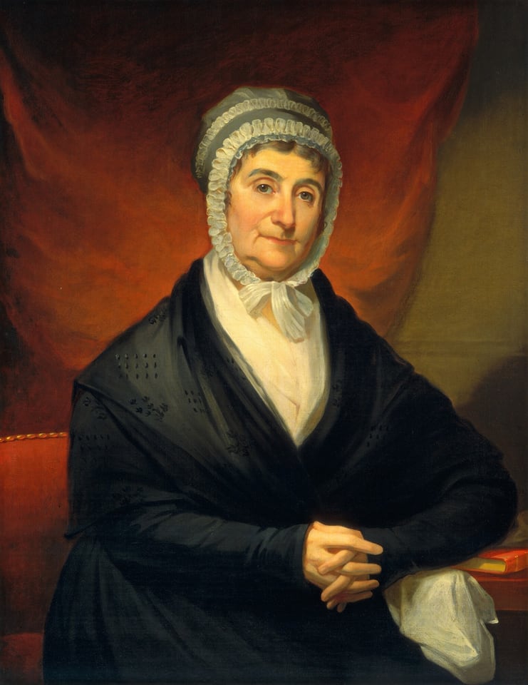 The image shows the upper half of a seated woman facing forward. She has her hands clasped together in front of her. The woman appears calm with visible signs of age, including a few wrinkles on her face. She wears a white bonnet with a frilled edge tied under her chin, a dark shawl over a white high-collared blouse, and sits on a red chair with a gold trim. A red book and a handkerchief are on the surface next to her. The background transitions from deep red to brown tones.