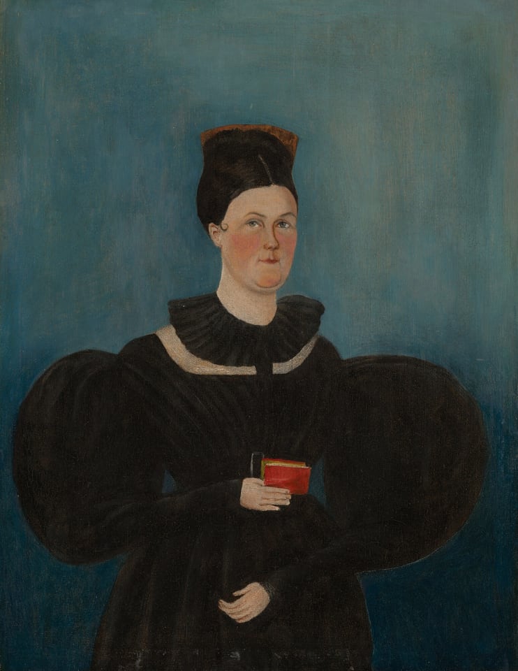 The image shows a woman standing upright with a small red book in her right hand. She has a fair complexion, neutral expression, slightly arched eyebrows, and small, rounded lips. Her dark brown hair is styled in an updo with a black accessory. She wears a high-necked dark dress with puffed sleeves and a white collar. The background is plain blue. The woman is not wearing any visible jewelry, and the setting is minimalistic with only the book present.