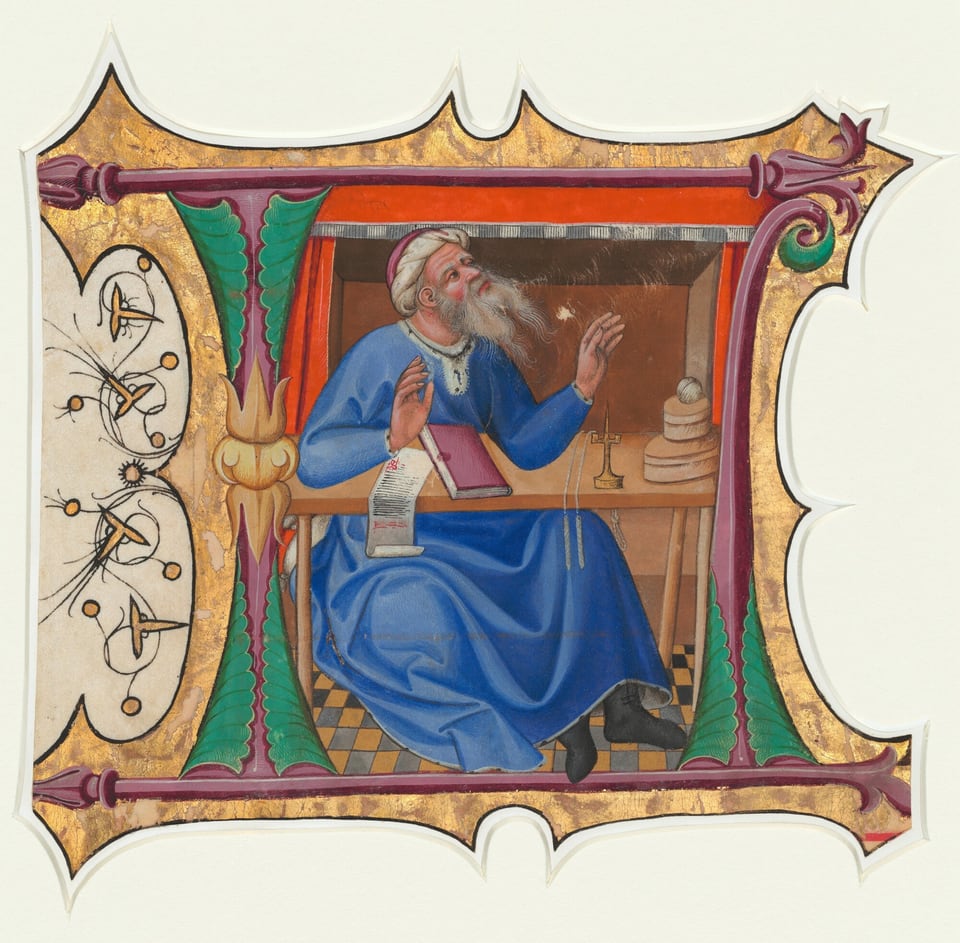 The image shows an elderly man seated at a table. He is turned slightly to the side, with his right hand raised as if in conversation. The man has a long, white beard and fair complexion, wearing a blue robe with white trim. He wears a white cap and holds a pink book in his left hand. A scroll of paper hangs off the table, which also holds a globe, balance scale, and round objects. The background has a wooden panel, and the scene is framed by an ornate border with gold, green, and purple colors, typical of medieval manuscripts.